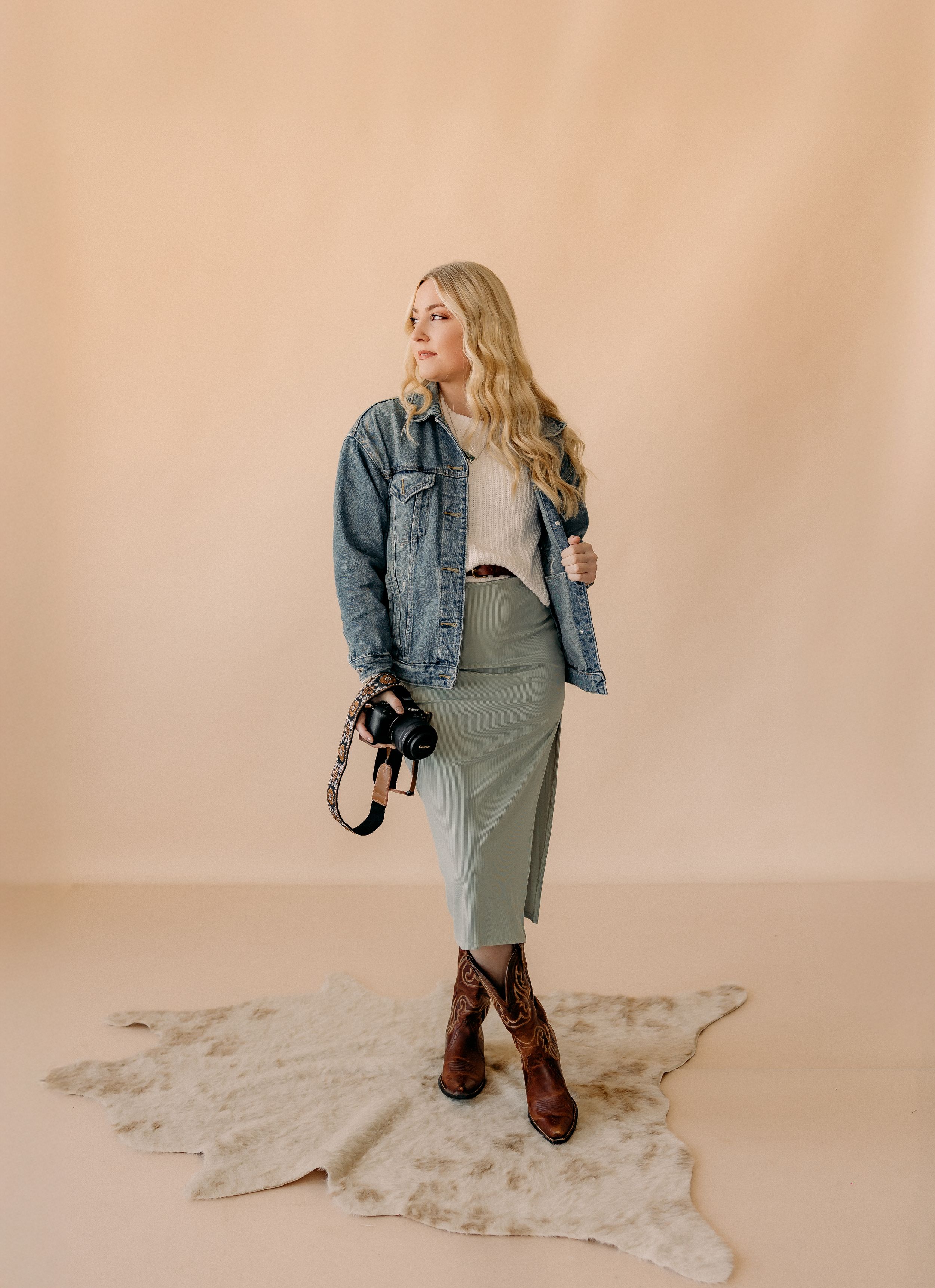 Person in denim jacket and dress poses on white rug against beige studio backdrop.
