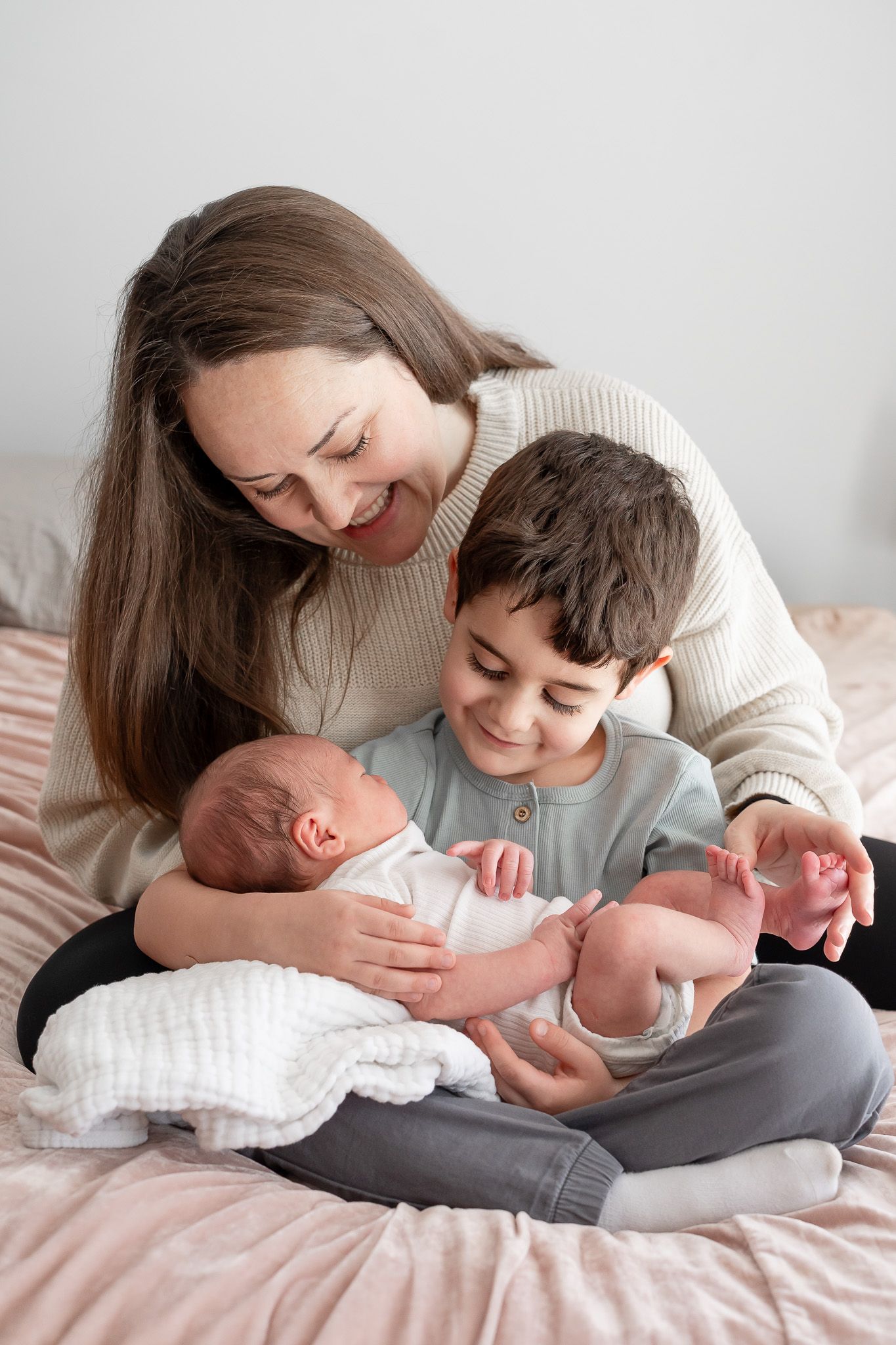 A tender family moment on a bed as siblings welcome their new baby sibling in matching neutral colored sweaters.