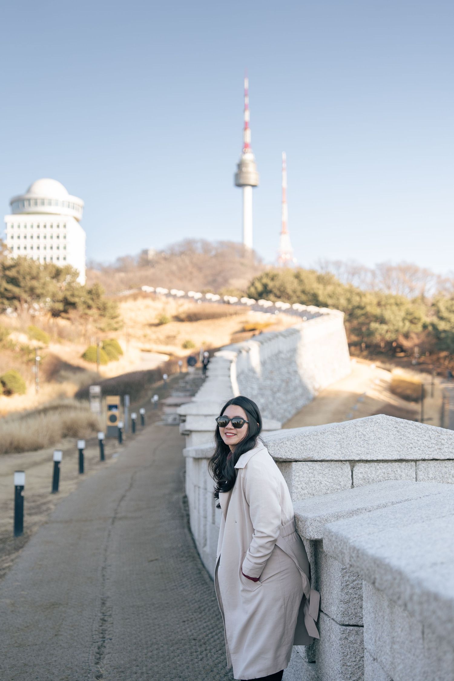 Scenic walkway along a hilltop overlook with white walls and communication towers in the distance on a sunny day.
