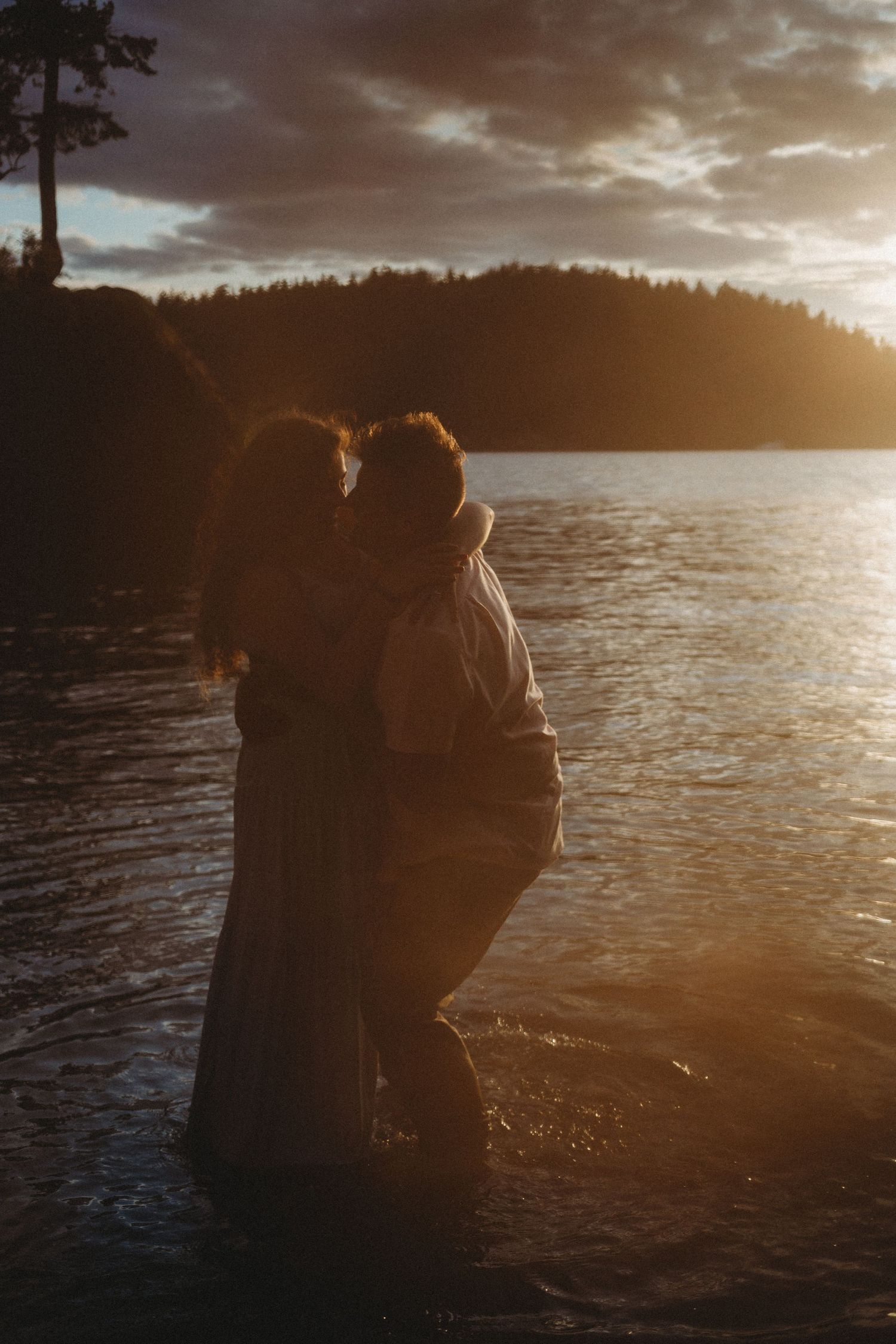 An engaged couple lean in close, noses almost touching in beautiful golden sunset light by the ocean