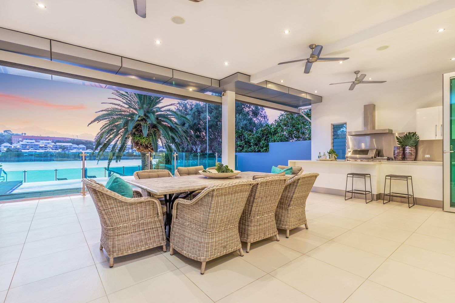 Spacious alfresco dining area with wicker chairs, ceiling fan, outdoor kitchen, and breathtaking waterfront sunset views.