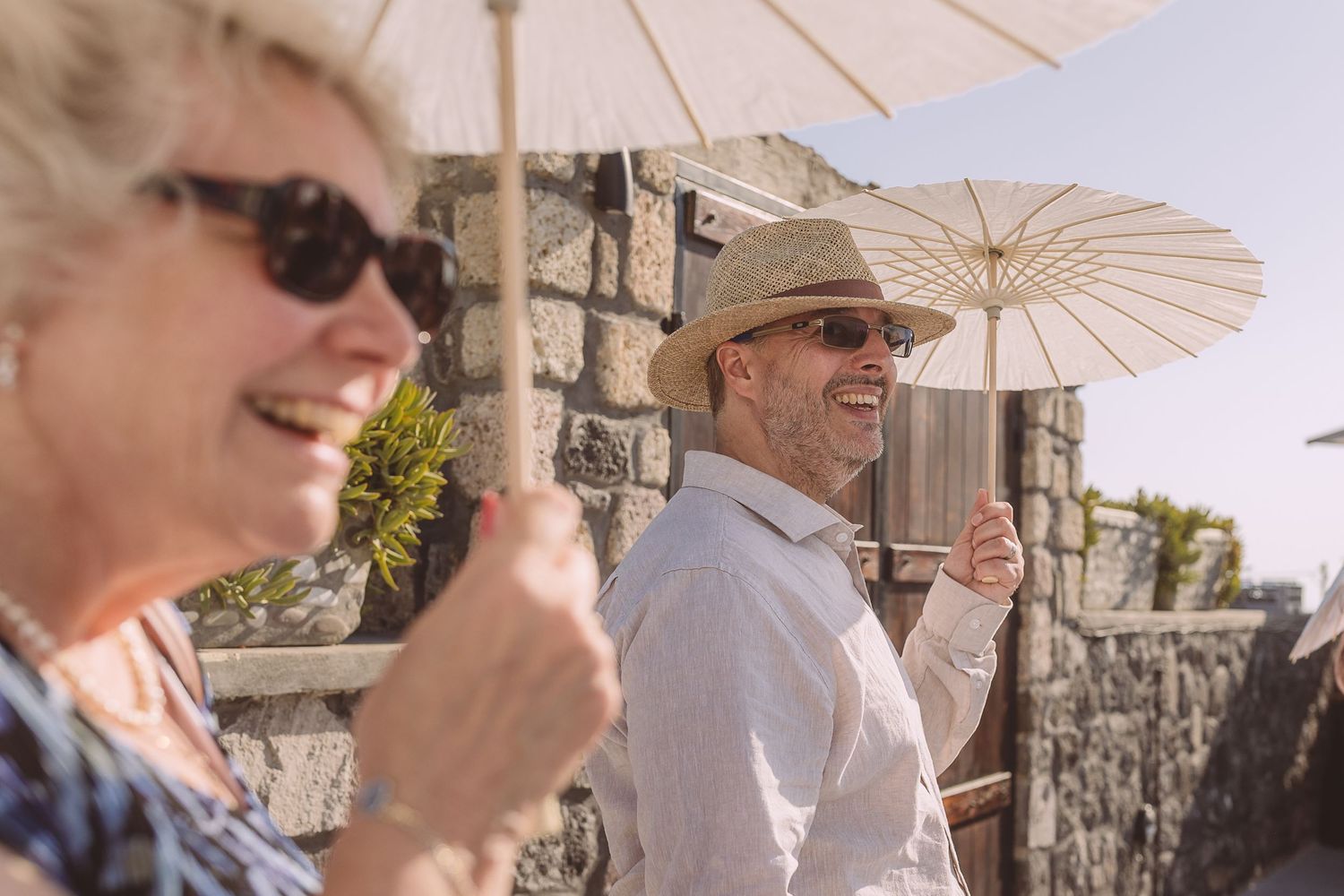 Two wedding guests at El Viento smile with parasols along a stone wall on a bright sunny day in Santorini.