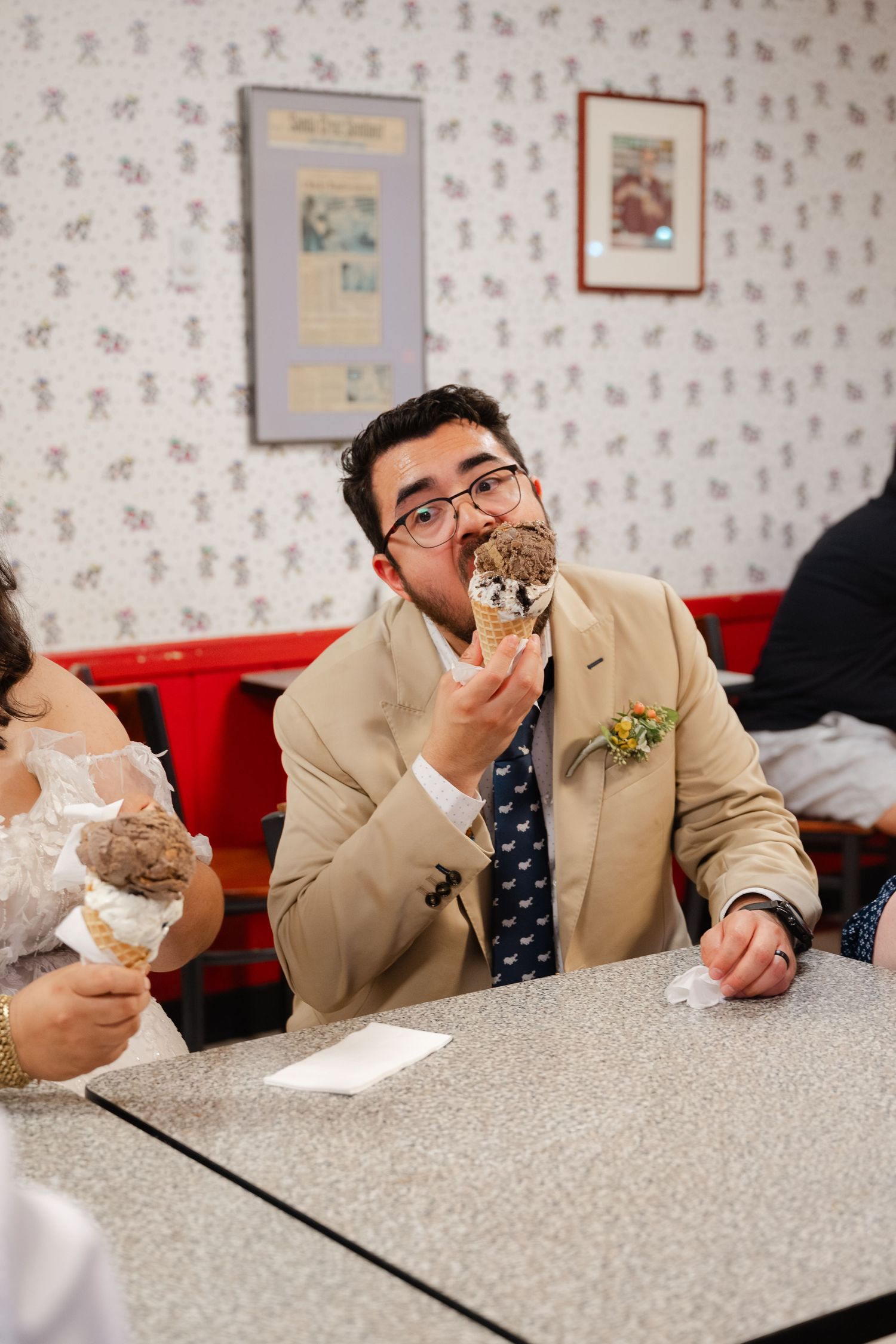 A person in a beige suit and tie eats a sandwich while sitting at a diner counter.