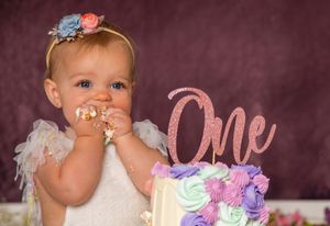 Baby in white dress with floral headband celebrates first birthday with colorful cake and rose gold 'One' topper.