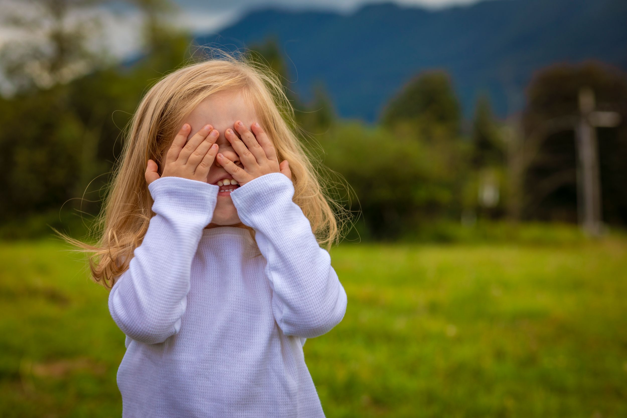 A person in a white sweater covers their face with hands while standing in a grassy field with mountains in background.