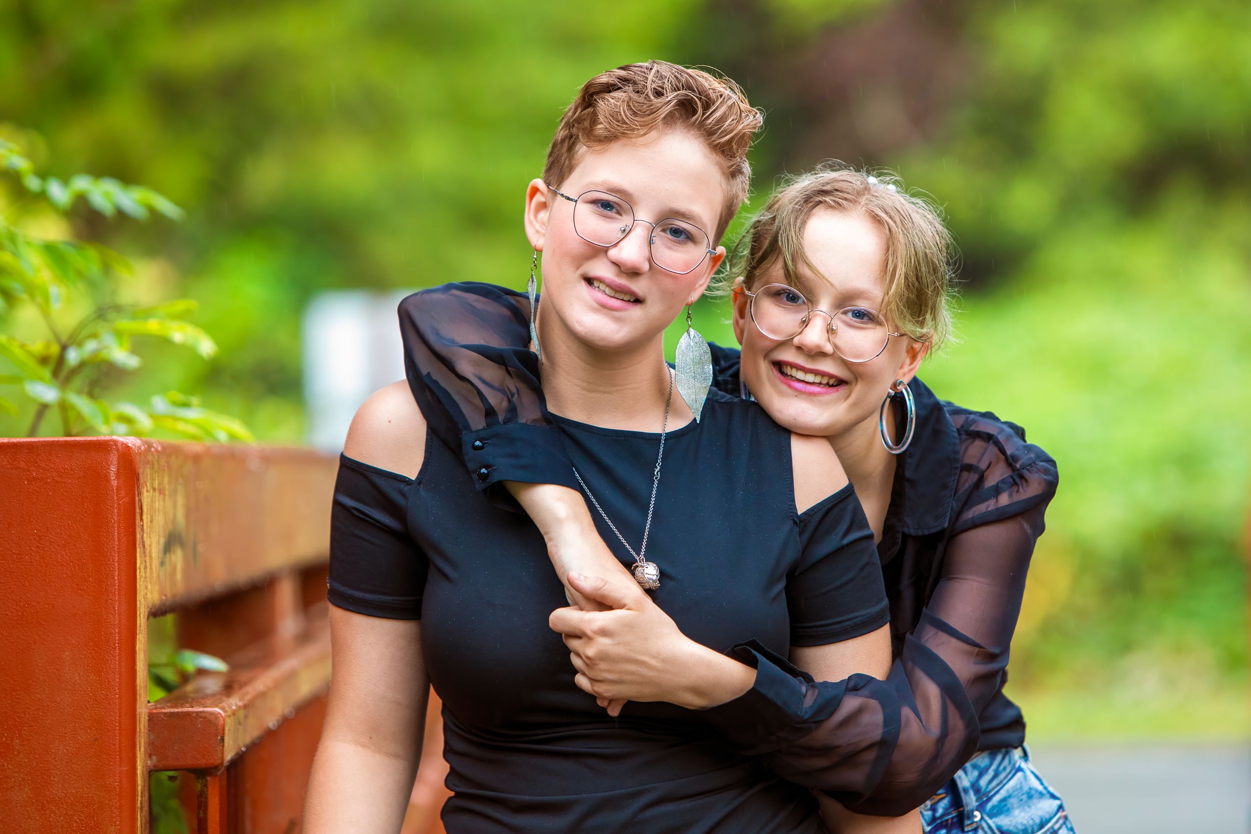 Two people in black outfits smile and embrace while sitting on a wooden bench outdoors in a green setting.