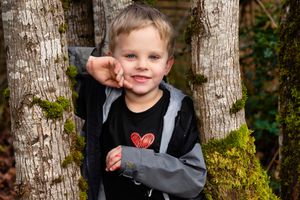 A child wearing a black jacket poses between two birch tree trunks in an outdoor nature setting.