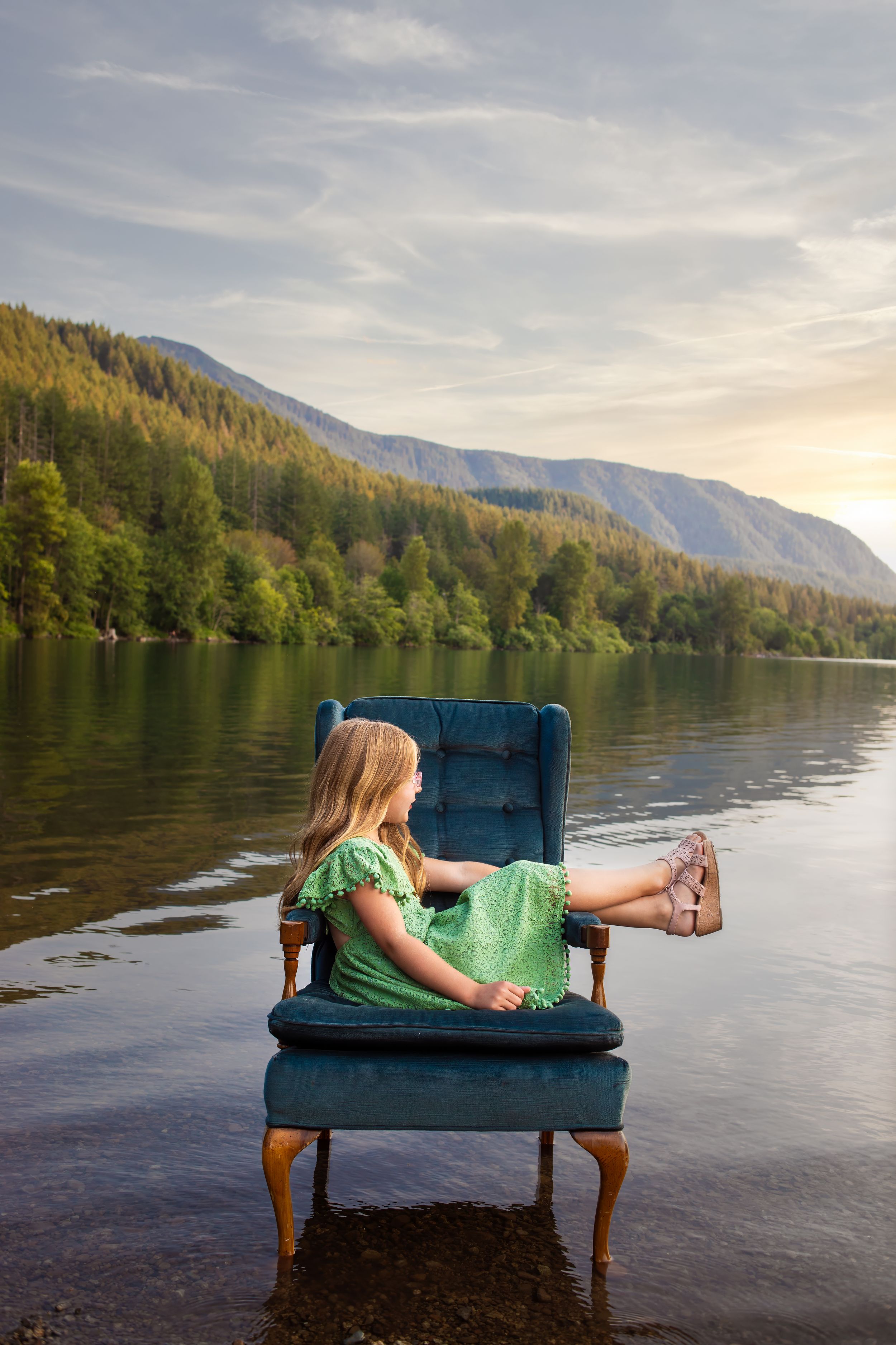 A teal velvet couch sits on a tranquil lake surrounded by mountains at sunset, creating a dreamlike scene.