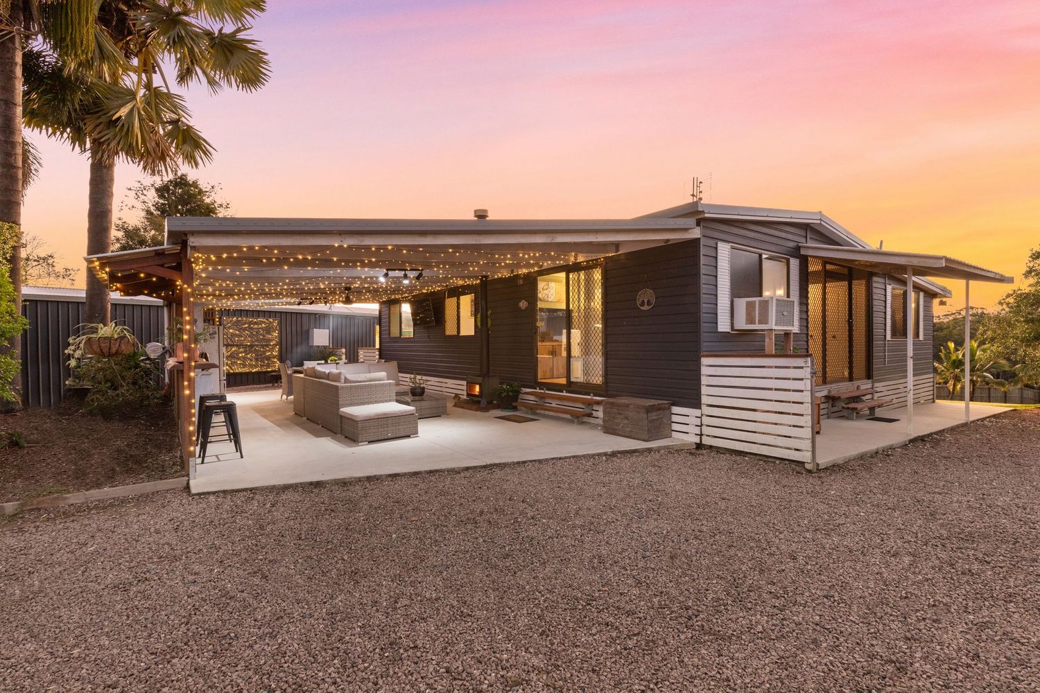 Contemporary dark-clad home exterior with covered pergola, outdoor seating, and gravel landscaping at sunset.