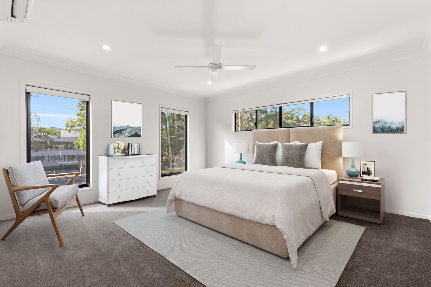 Bright master bedroom with carpet, ceiling fan, large windows, neutral bedding, timber bedside tables, and white dresser.