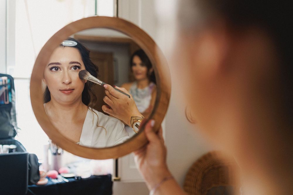 Bride having makeup applied while looking into a hand mirror during wedding preparations.