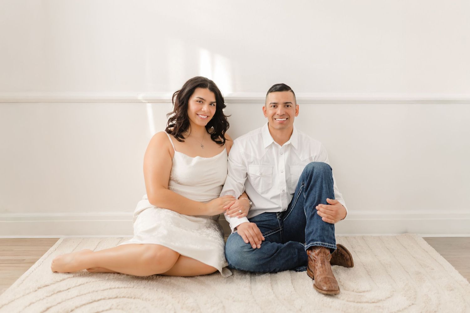 Couple lounging together on white floor during minimalist indoor photoshoot with natural lighting.