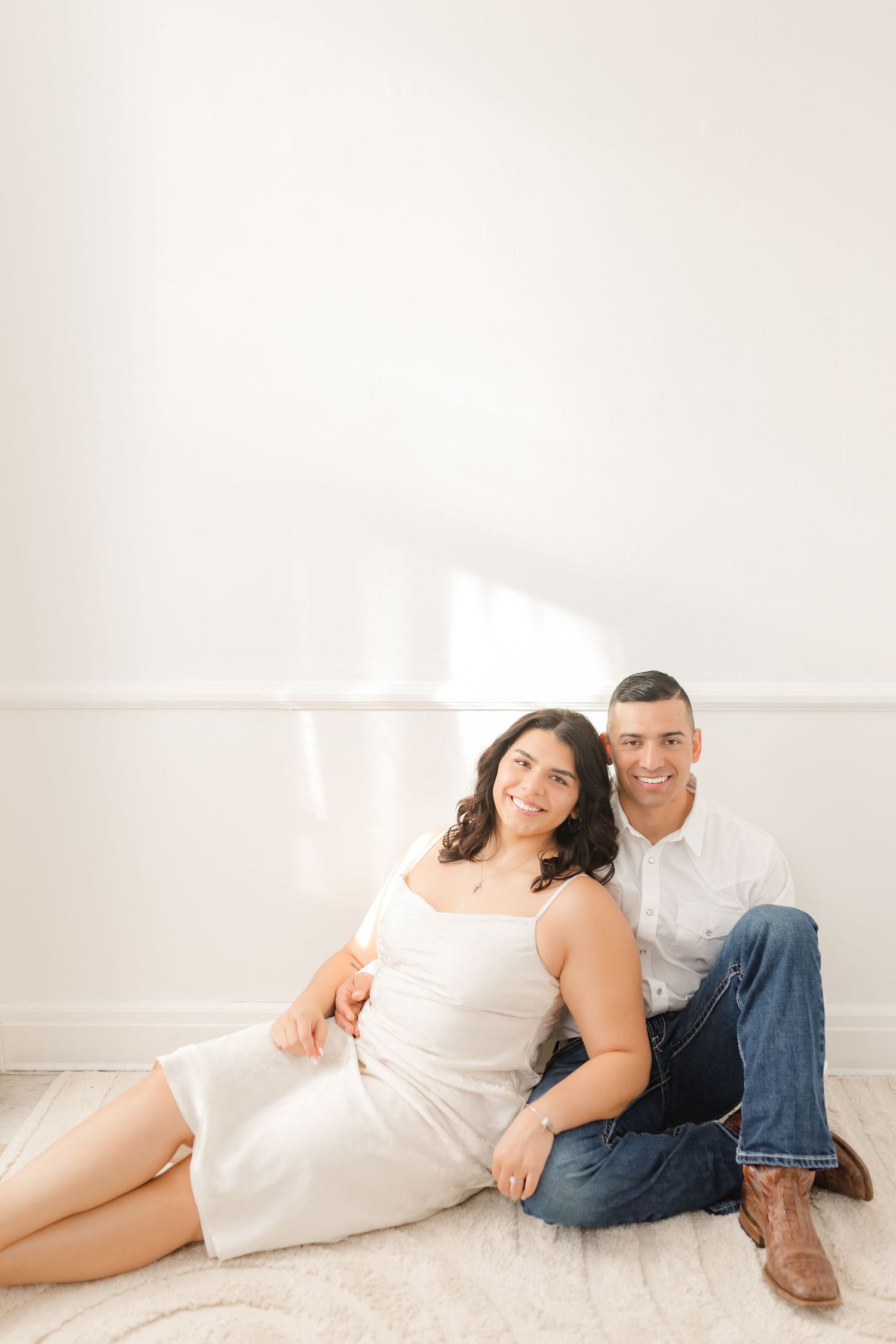 A loving couple in casual attire pose together on the floor in a bright, minimalist setting.