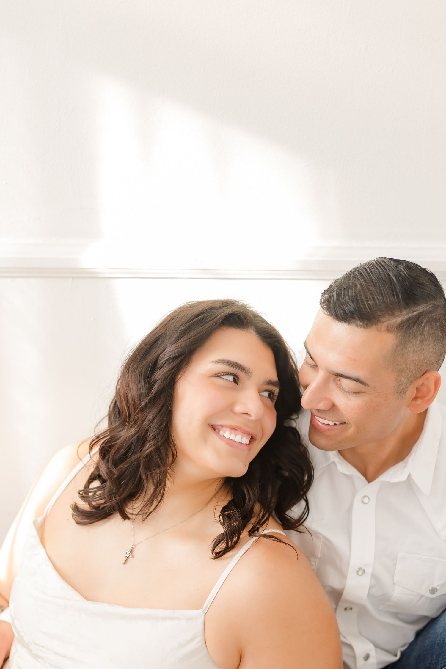 A couple shares a tender moment together while smiling and embracing in a bright, naturally lit room.