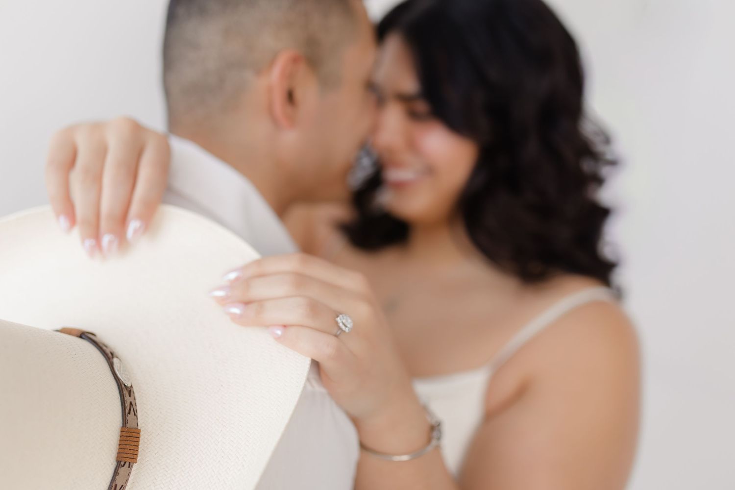 Couple in white outfits embracing in a romantic engagement photo session against neutral background.