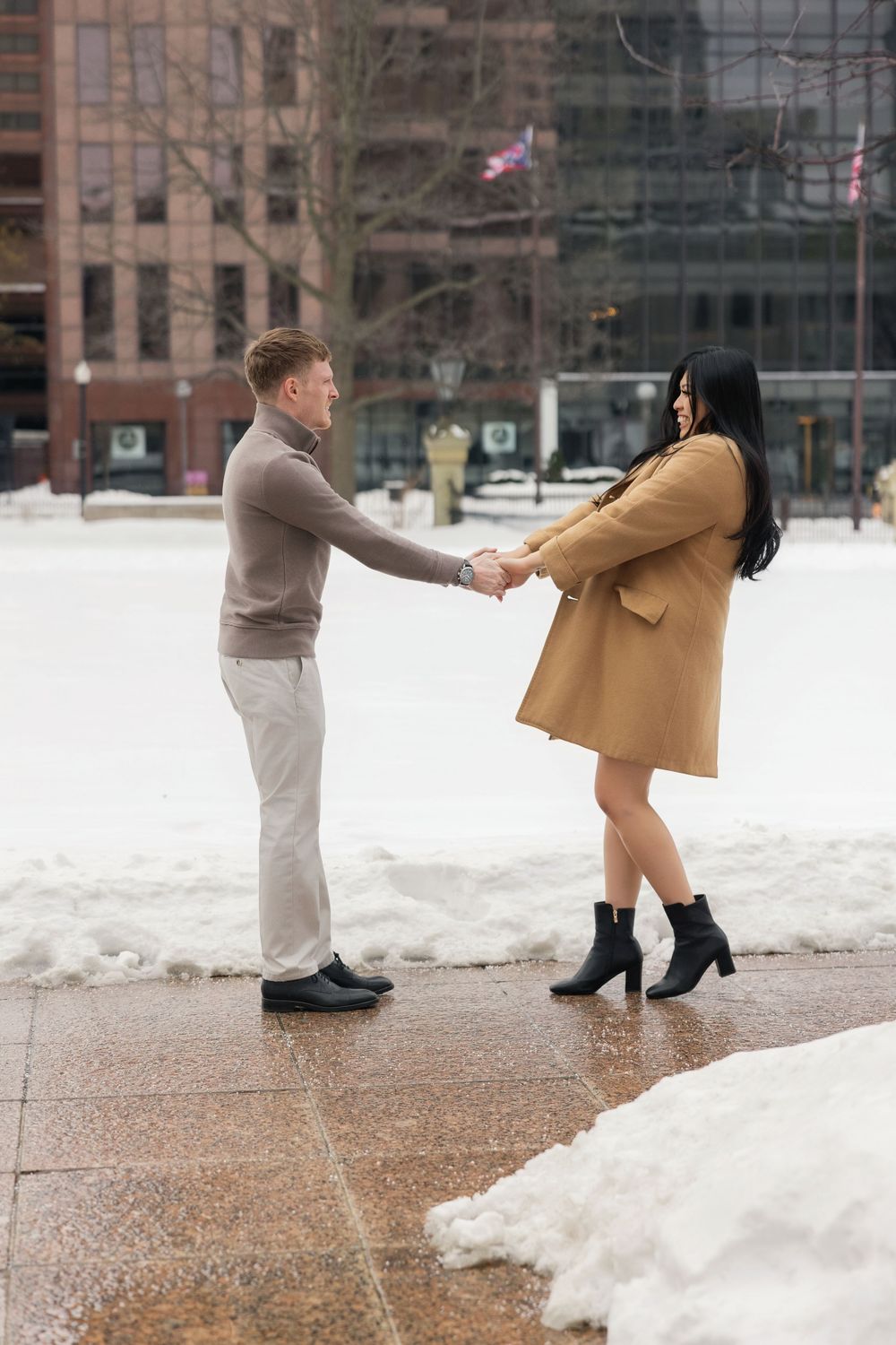 A couple holds hands while standing in a snowy urban plaza with American flags visible in the background.