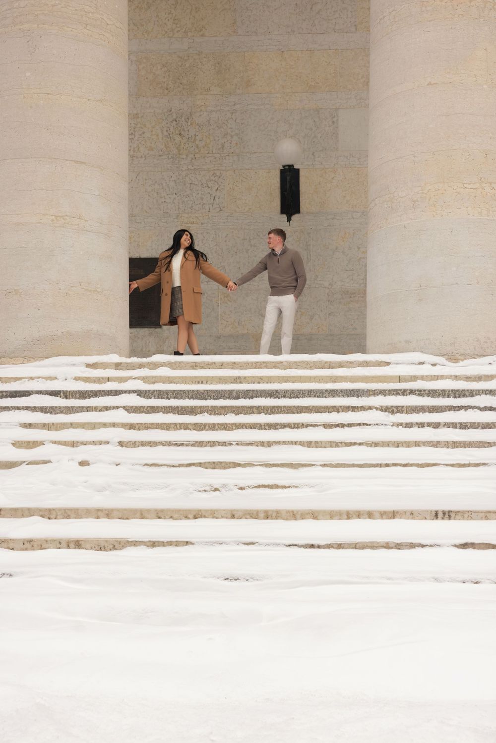 Two people stand on white stone steps in front of a light-colored stone wall in a minimalist architectural setting.