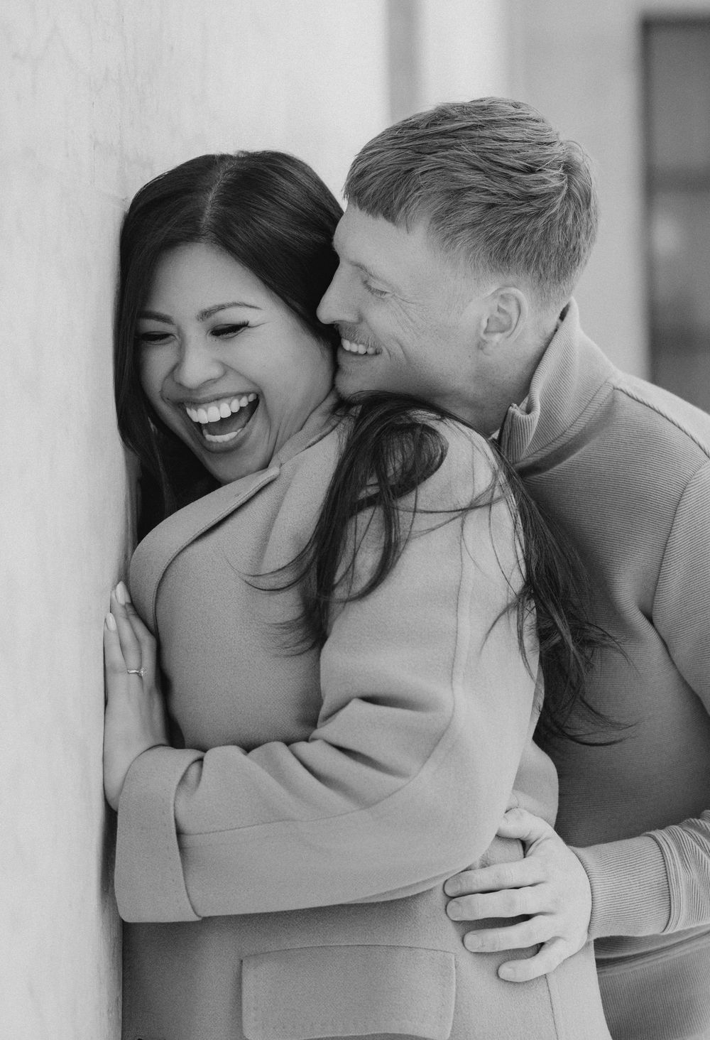 A joyful black and white portrait of a couple laughing and embracing against a wall.