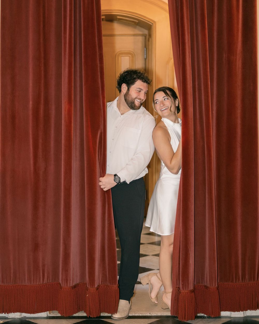 A couple shares a playful moment behind red velvet curtains.