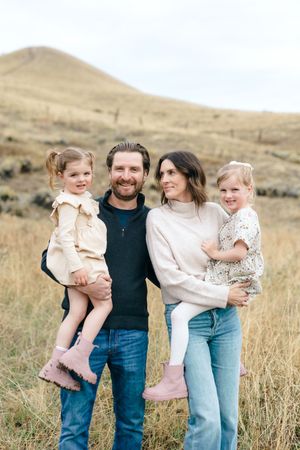 Family wearing casual clothing poses together in golden grass field with rolling hills in background.