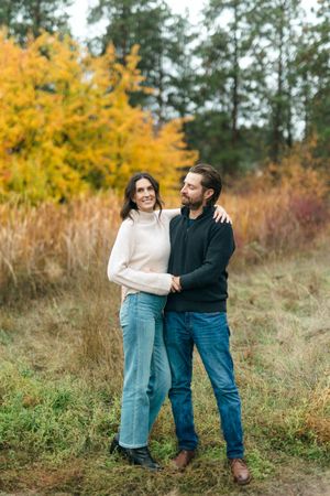 A couple in casual sweaters embraces while standing on a nature trail with golden autumn foliage in the background.