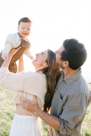 A loving family plays together outdoors in a sunlit field, lifting and embracing their small child with joy.