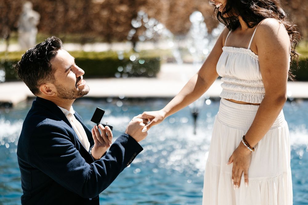 A romantic proposal scene by a fountain with someone in a navy suit kneeling before someone in a white dress.