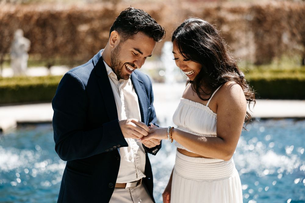 Couple sharing an emotional moment together by a fountain in formal attire on a sunny day.
