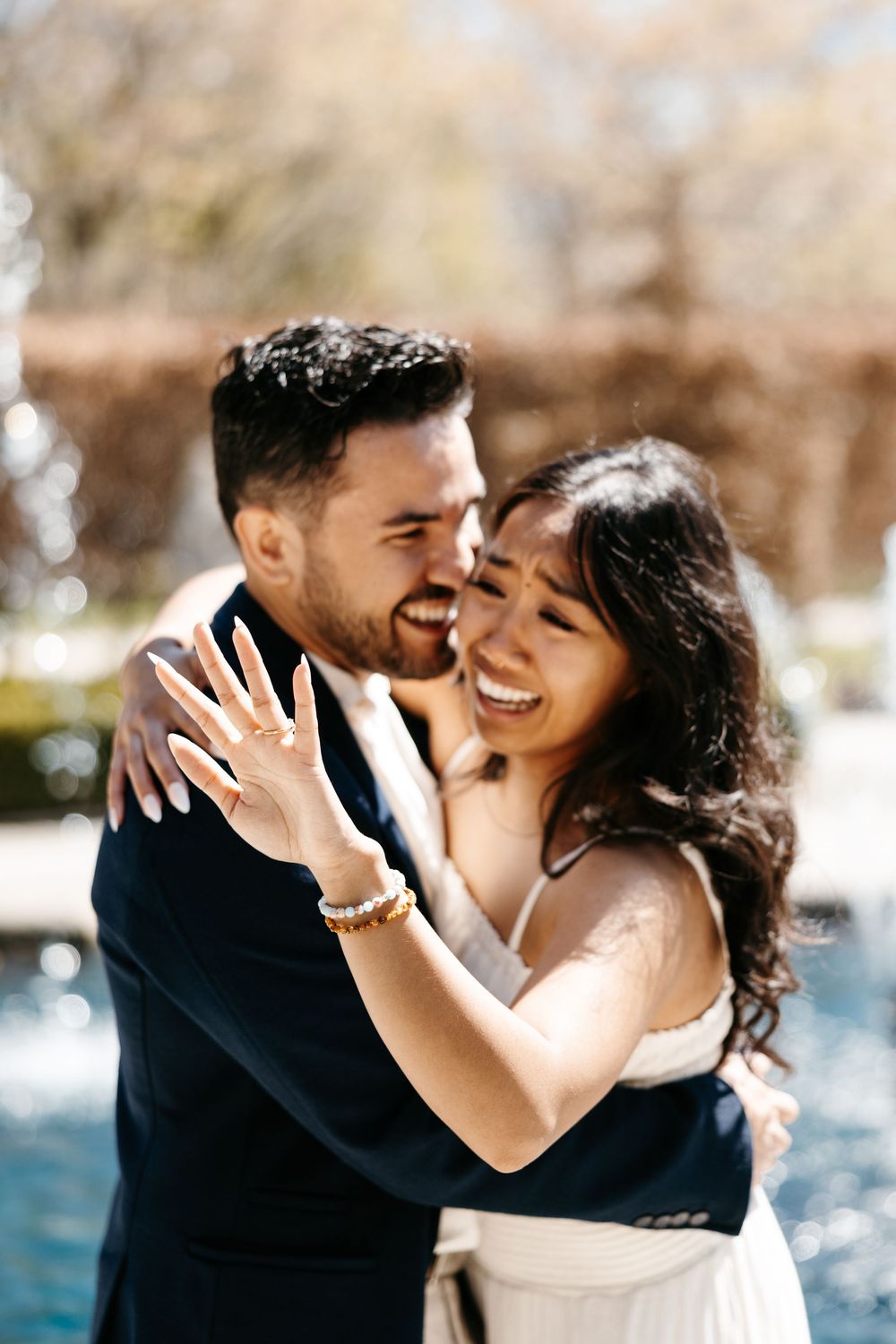 Happy couple embraces and laughs while showing off engagement ring by pool in outdoor setting.