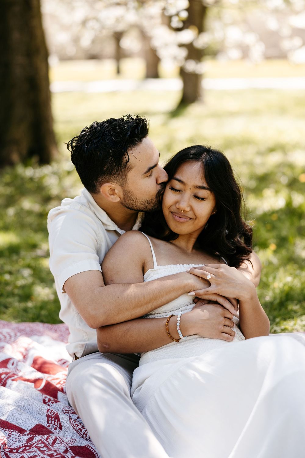 A romantic couple embraces on a blanket in a sunny park during an outdoor photo session.