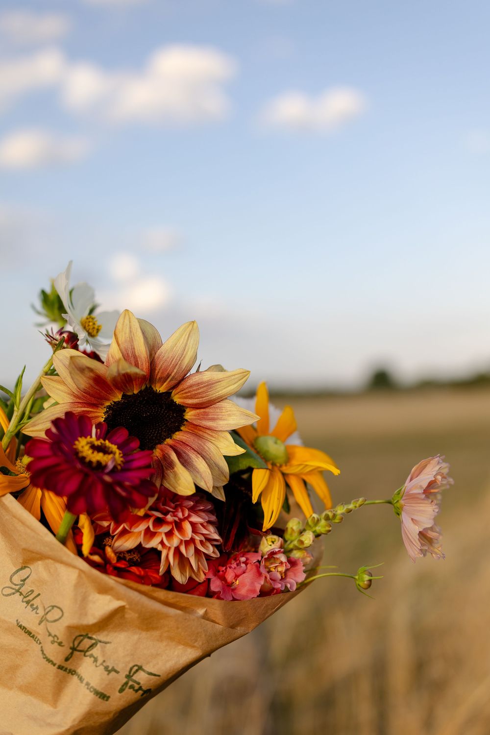A bouquet of vibrant sunflowers and wildflowers wrapped in brown paper sits against a golden wheat field at sunset.