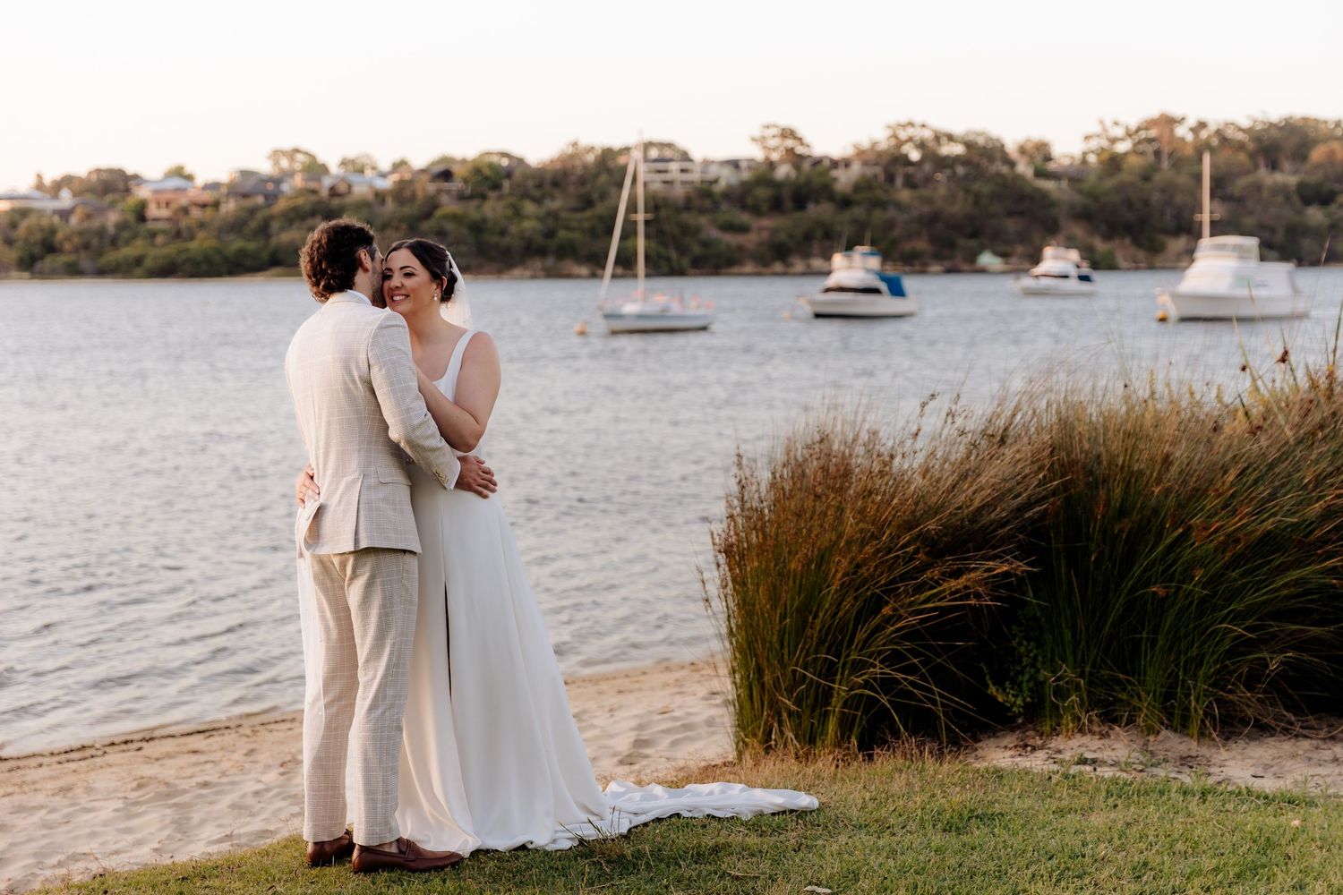 A couple shares an intimate moment by the waterfront with sailboats anchored in the harbor at sunset.