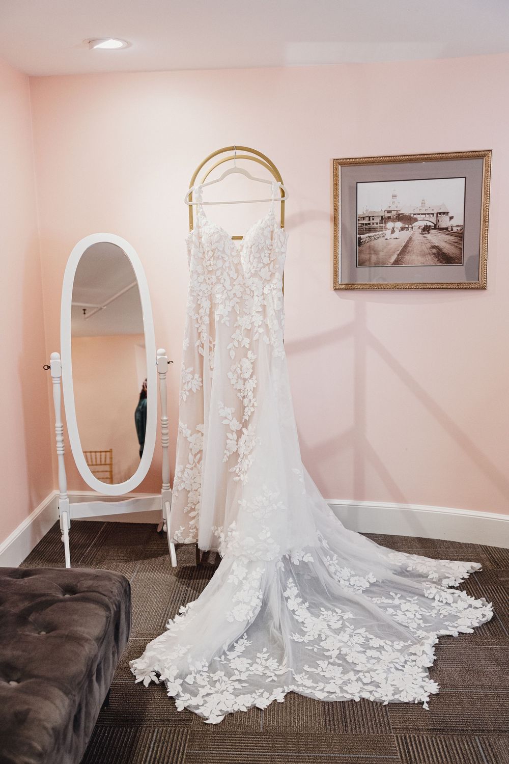 A stunning lace wedding dress with a long cathedral train displayed in a bridal suite with oval mirror and pink walls.