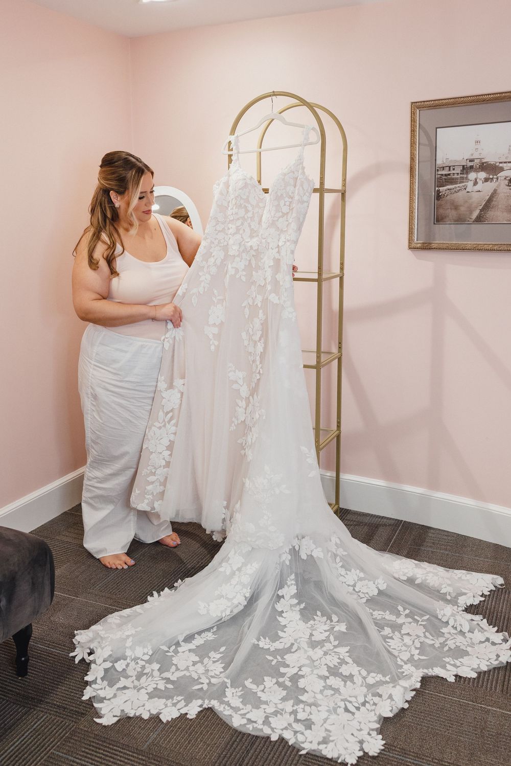 A wedding dress with delicate lace detailing and a long train is being arranged on dark carpeted flooring.
