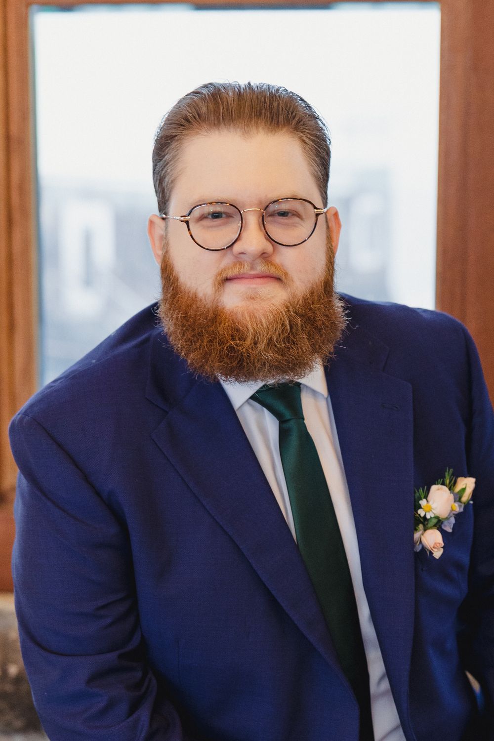 Groom in navy suit and green tie smiles while wearing round glasses and sporting a full red beard.