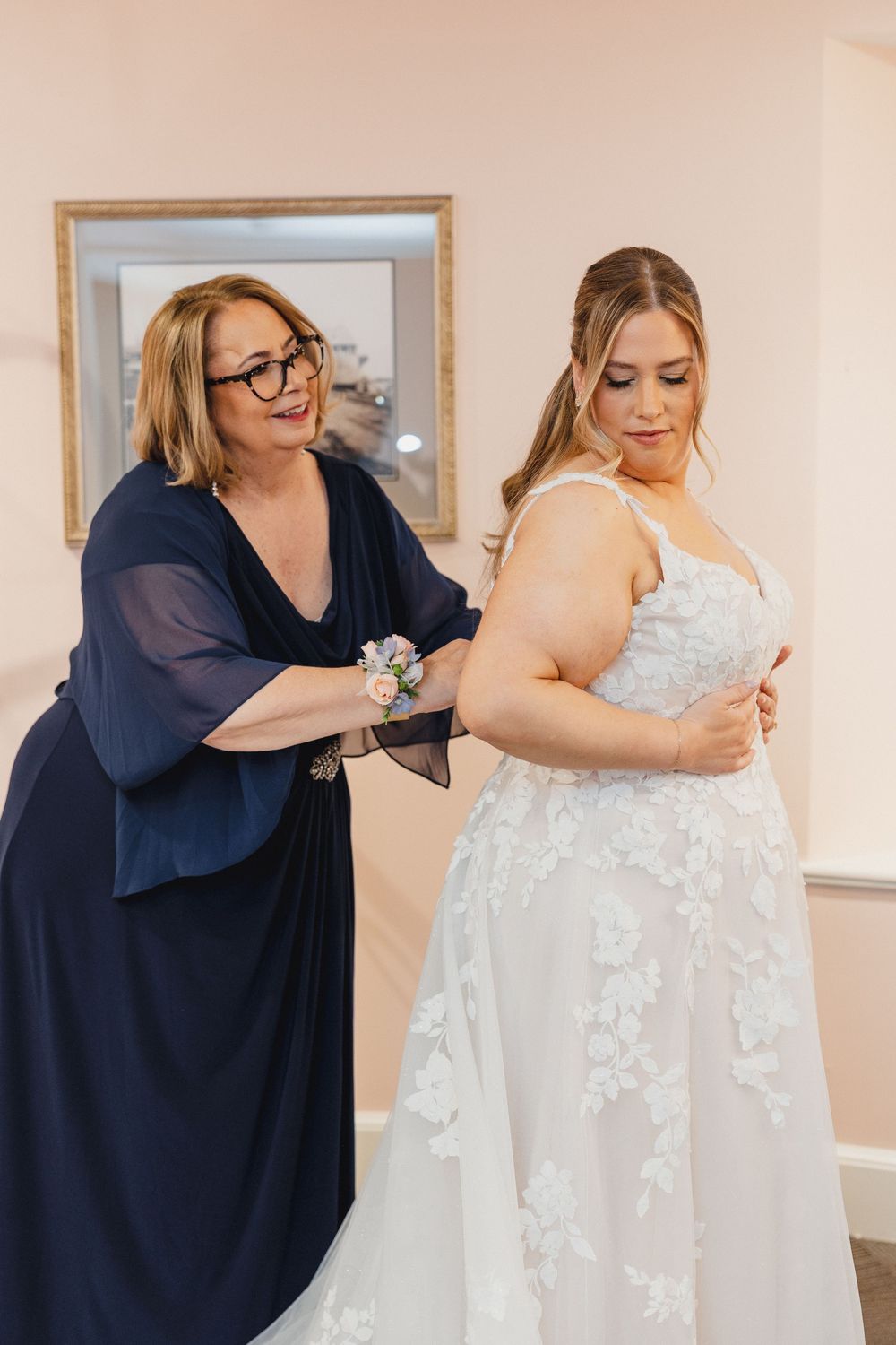 Wedding party helps bride with her white lace dress during preparation in a light-filled room.