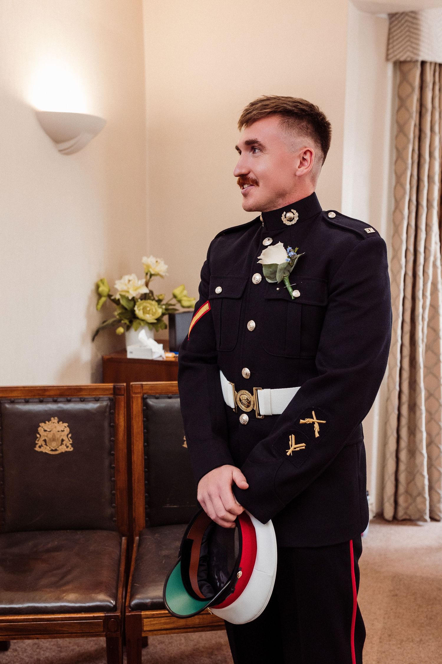 Military serviceman in dress uniform adjusts his formal attire while standing in an elegant interior setting.