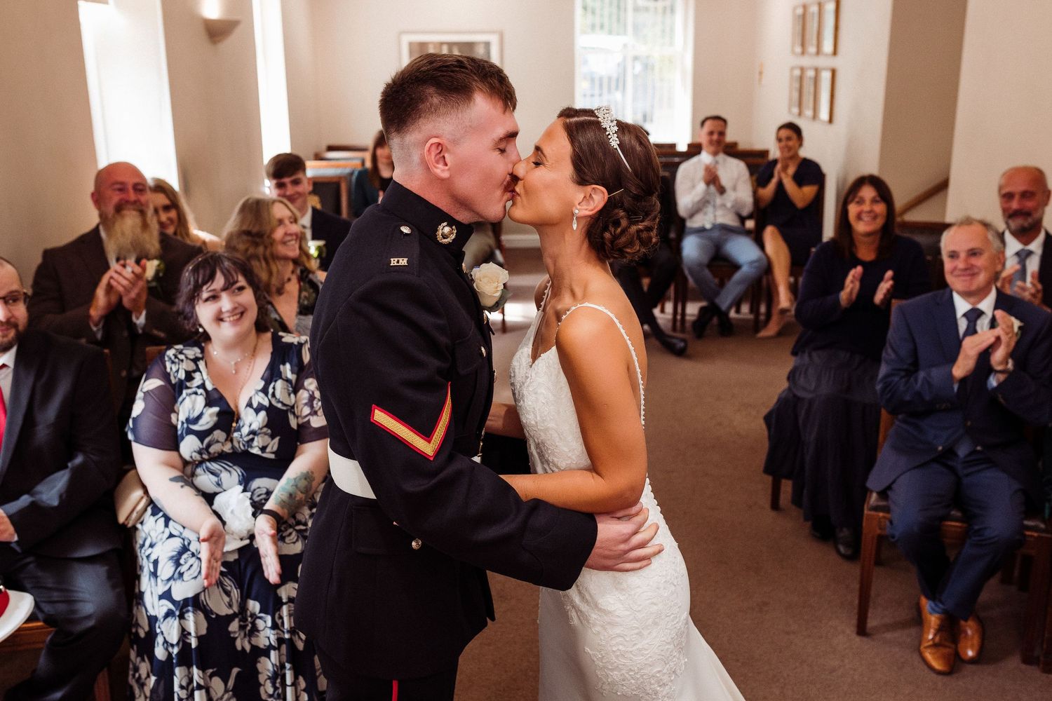 A romantic first kiss between newlyweds at their wedding ceremony as guests look on in a light-filled venue.