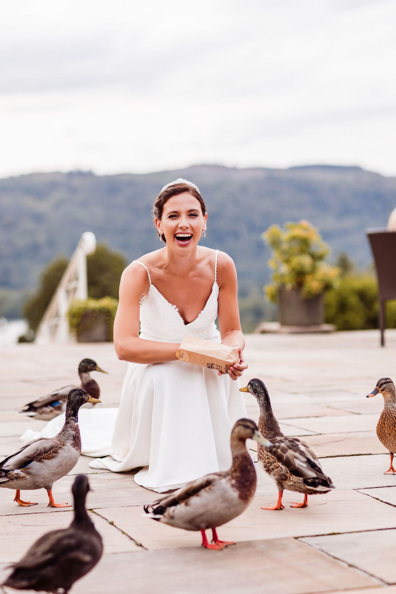 A person in a white dress laughs joyfully while surrounded by ducks on a stone patio with mountains in background.