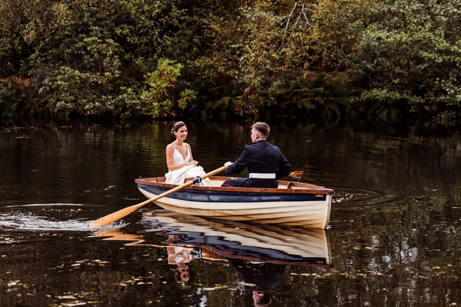 Couple rowing wooden boat on tranquil river surrounded by dark forest scenery.