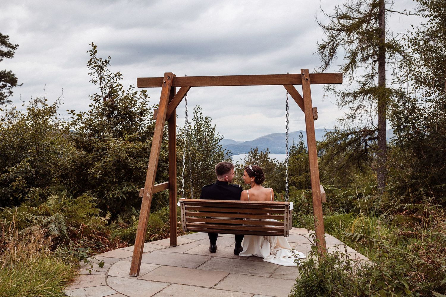 A wooden swing bench overlooks a scenic mountain vista on a stone patio surrounded by evergreen trees.