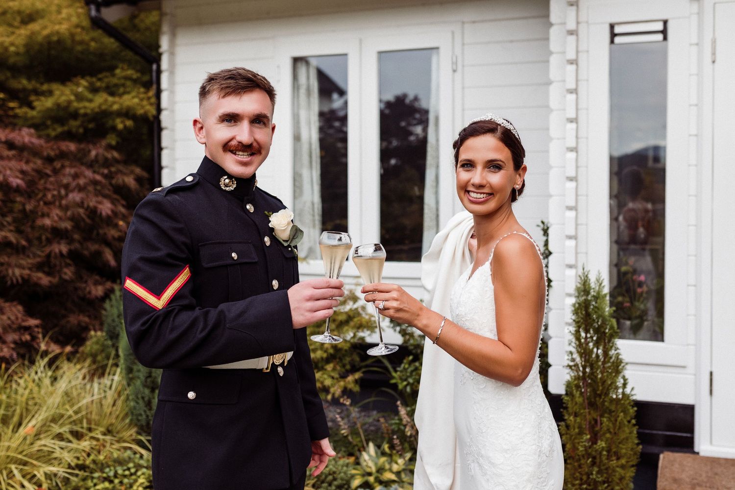 A couple in wedding attire shares a celebratory toast outside a white building with fall foliage in the background.