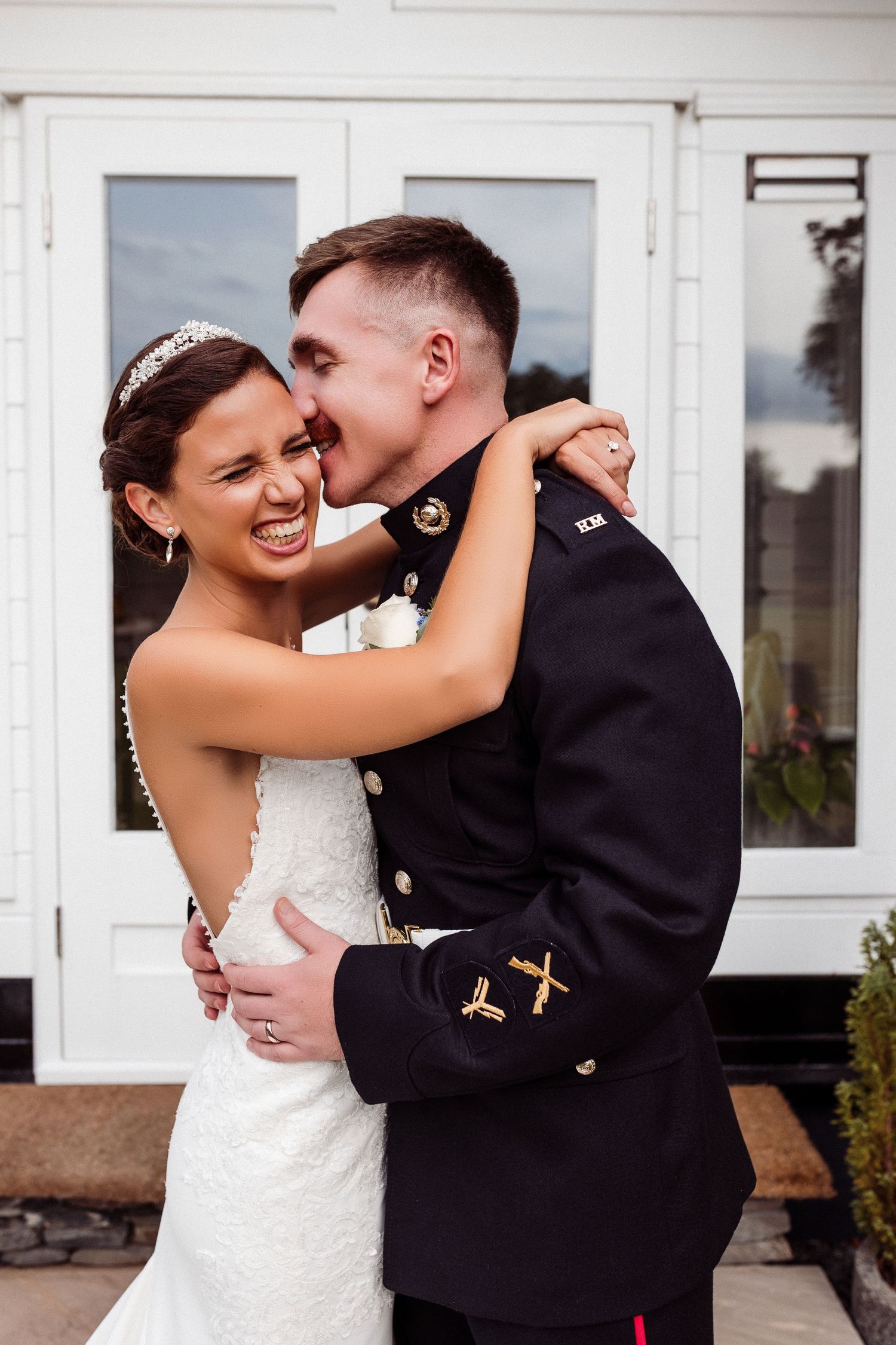 A couple shares intimate moments in wedding attire outside a white building with windows.