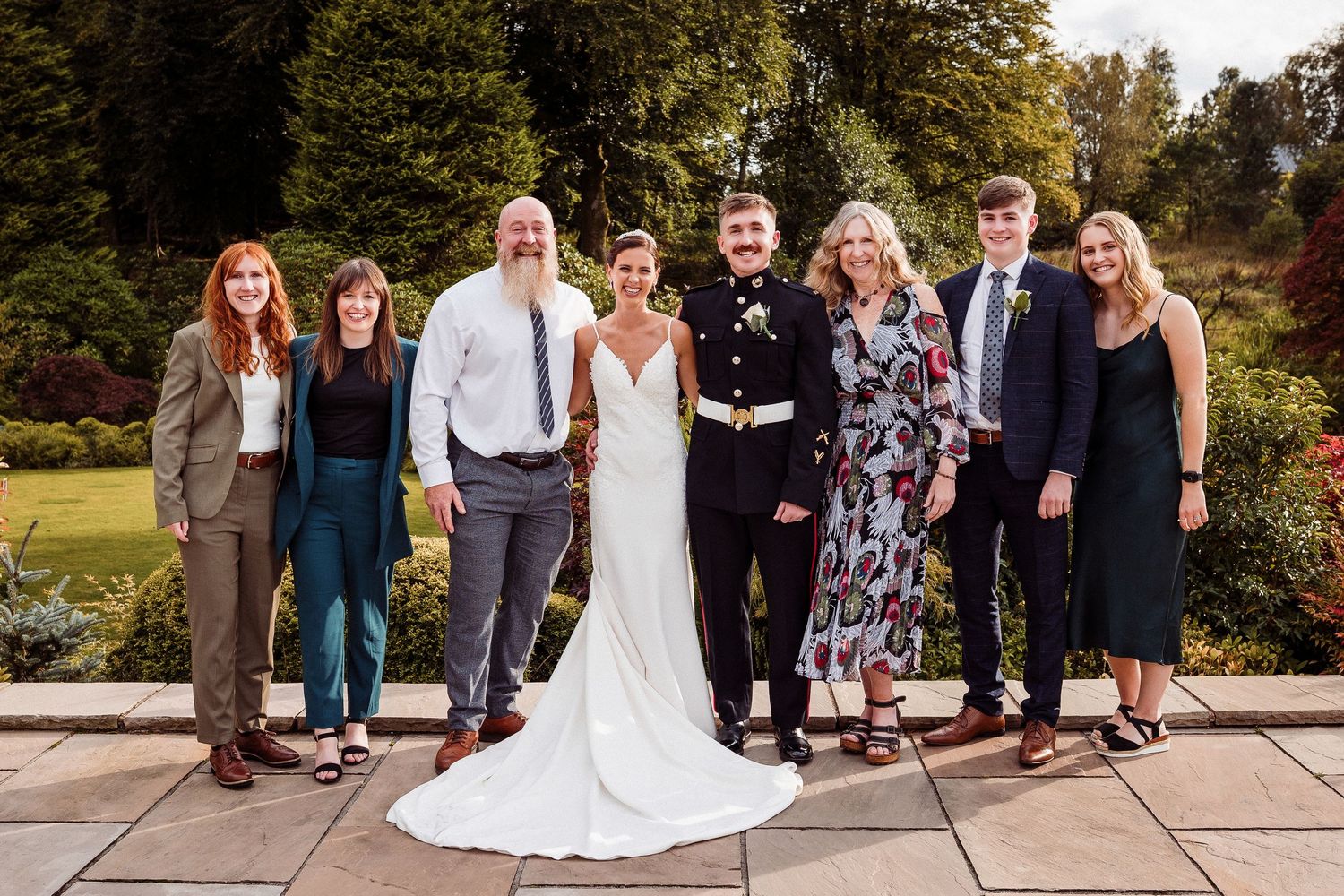 Wedding party and guests gather outdoors for group photos in a scenic garden setting during golden hour.