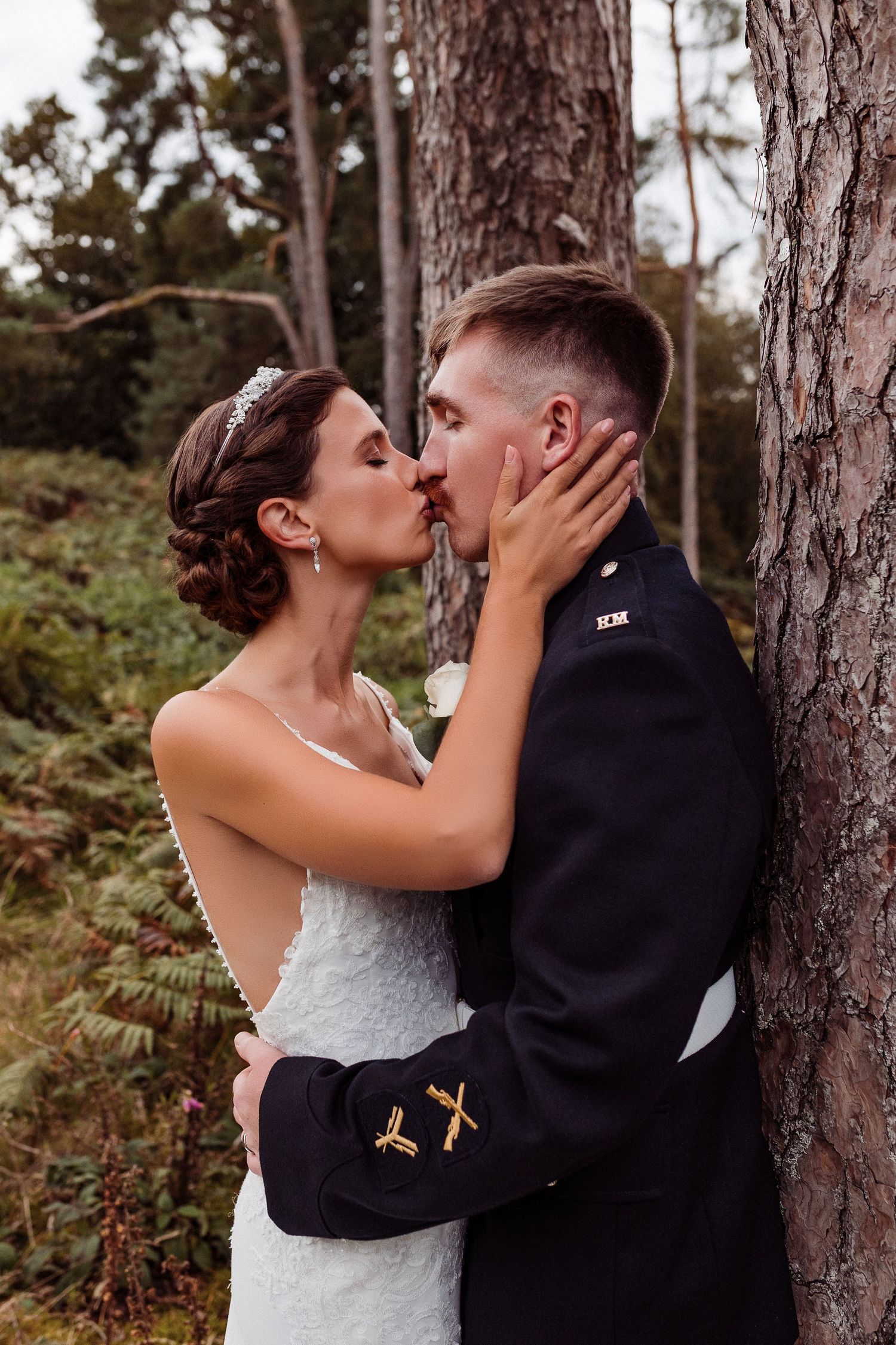 A romantic wedding couple shares an intimate kiss against a tree trunk in a wooded forest setting.
