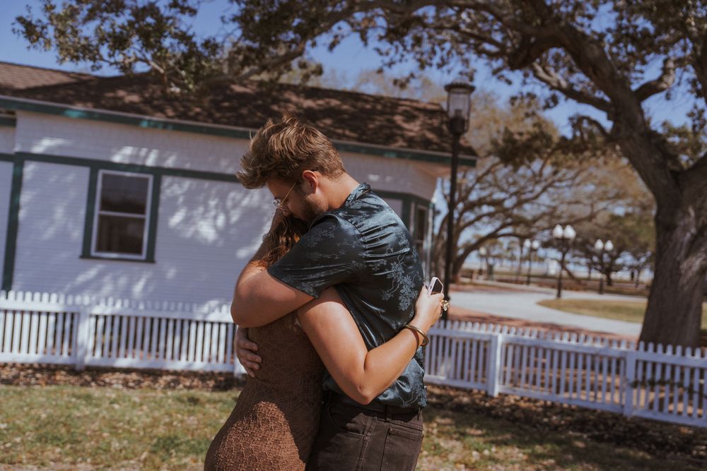 A romantic couple embraces and kisses under a tree in a sun-dappled outdoor setting.