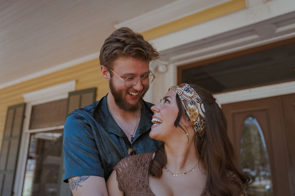 A couple shares a joyful moment together on a porch, with the person wearing a floral headband laughing.
