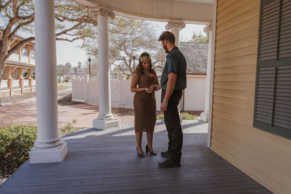 Two people stand in conversation on a historic porch with white columns and wooden floors in warm afternoon light.