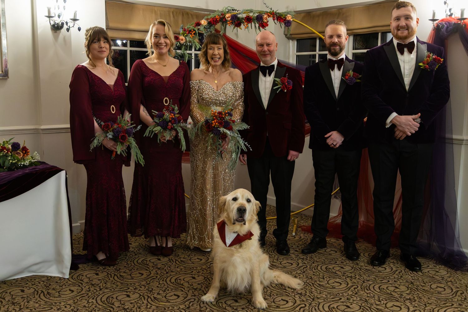 Wedding party in burgundy and black attire poses with a golden retriever wearing a floral collar at indoor venue.