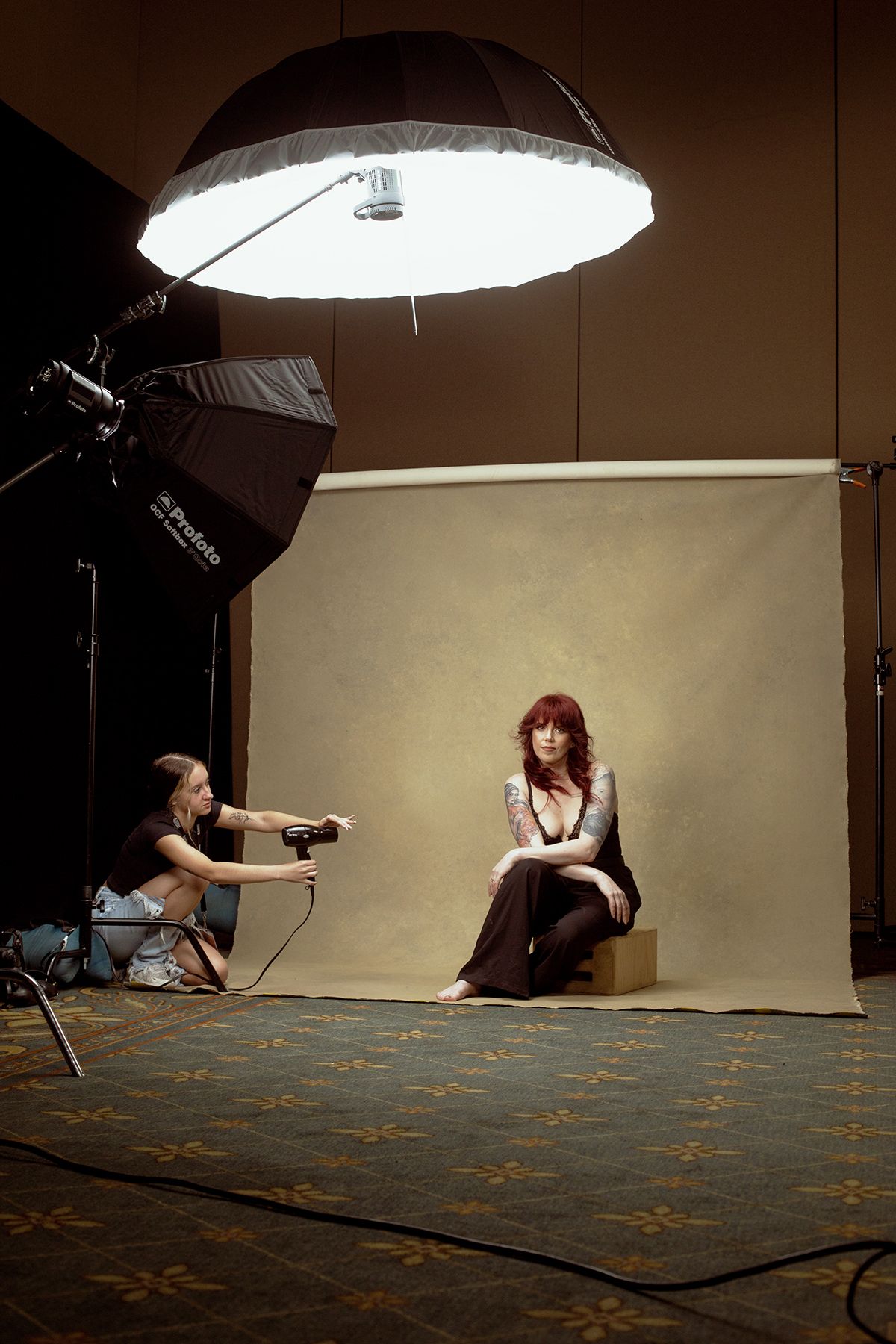 Professional photographer in black dress poses with camera against dark gray backdrop in artistic portrait series.